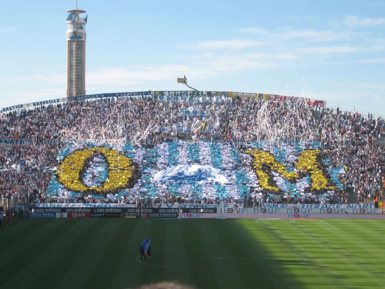 Cartulinas blancas, azules y amarillas formando un tifo en el Virage Nord del estadio Vélodrome